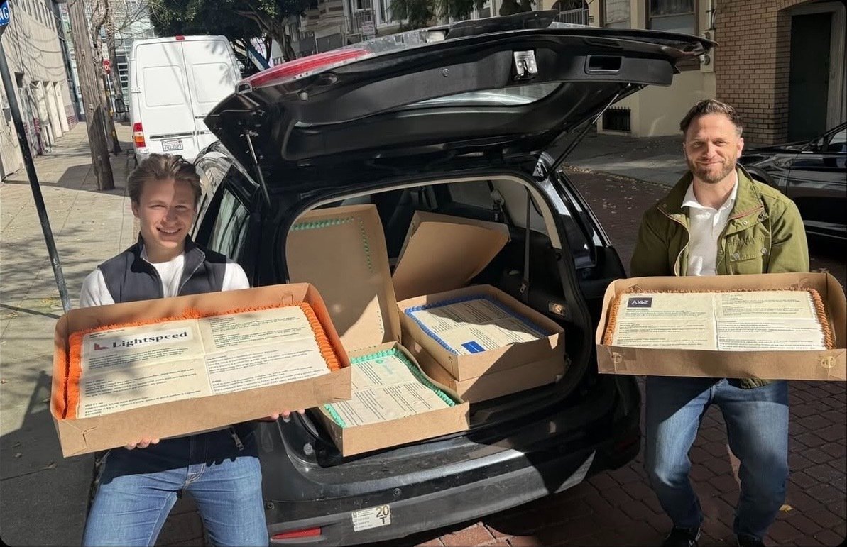 Two founders holding boxed pitch cakes beside a car, about to deliver them
