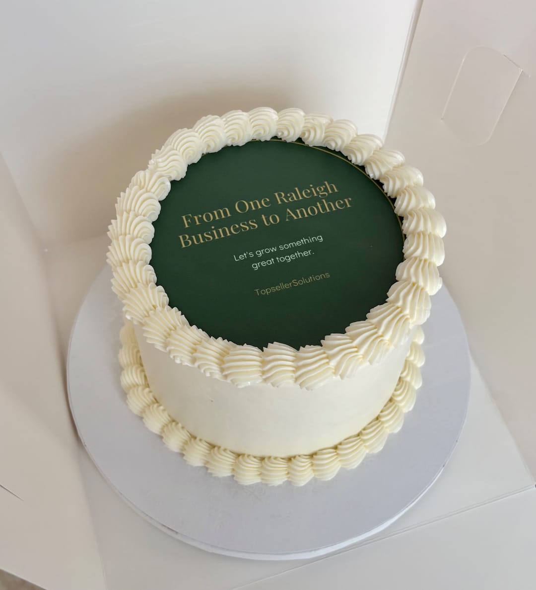 A small white cake with a green top reading From One Raleigh Business to Another